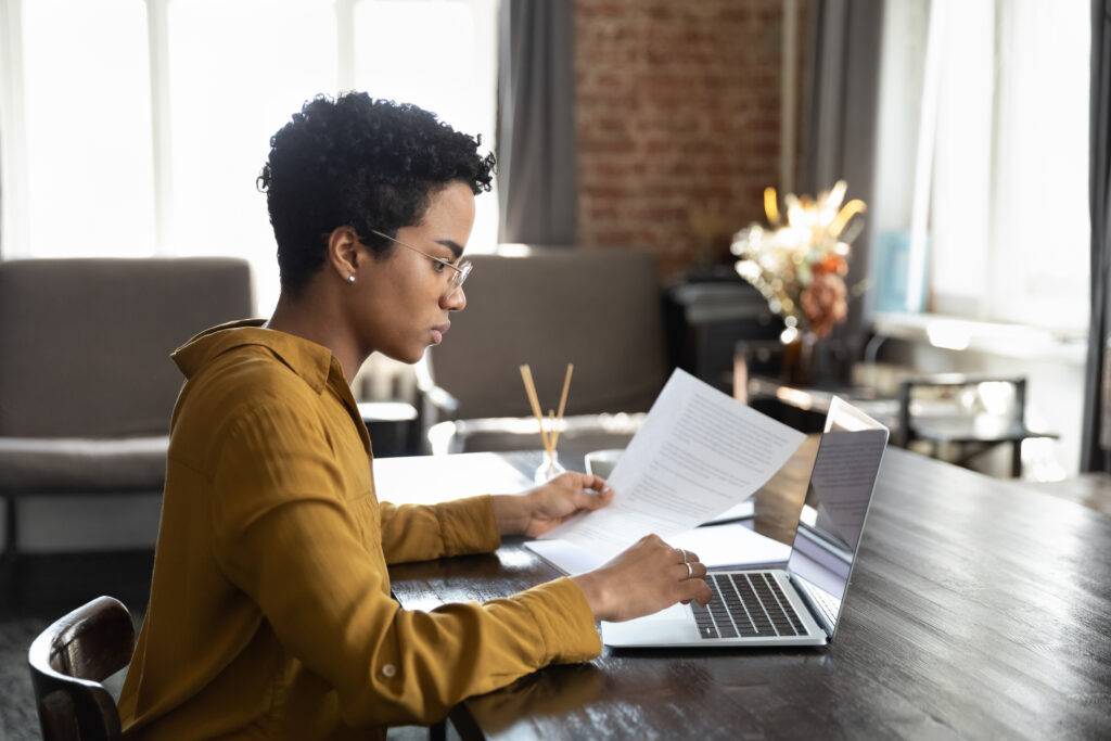 Focused serious young African American businesswoman manager employee in eyewear analyzing paper reports working on computer in modern office, preparing presentation or doing accounting paperwork.