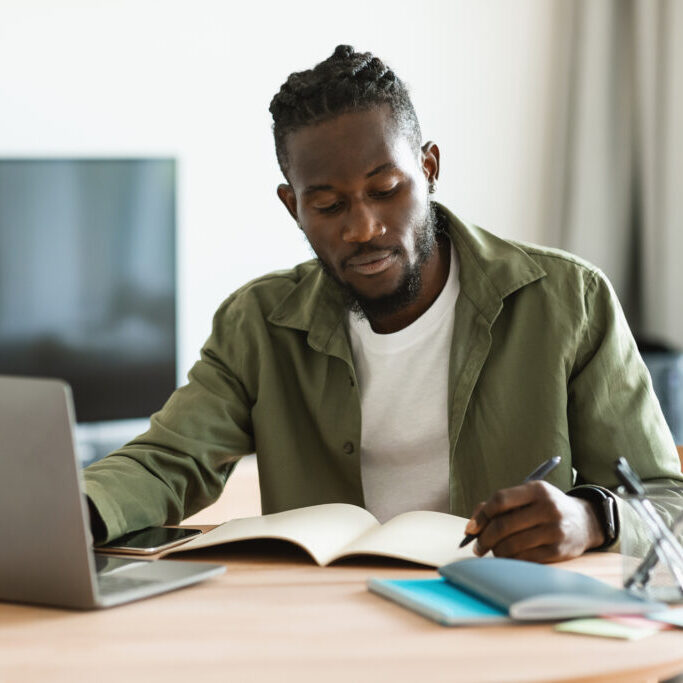 Focused african american man sitting at desk working on laptop and taking notes in notebook, studying online at home. Male watching webinar and using computer